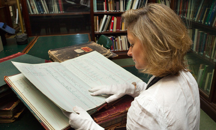 An image of a woman reading an old book in a library setting. She wears white cotton gloves to handle the object. 